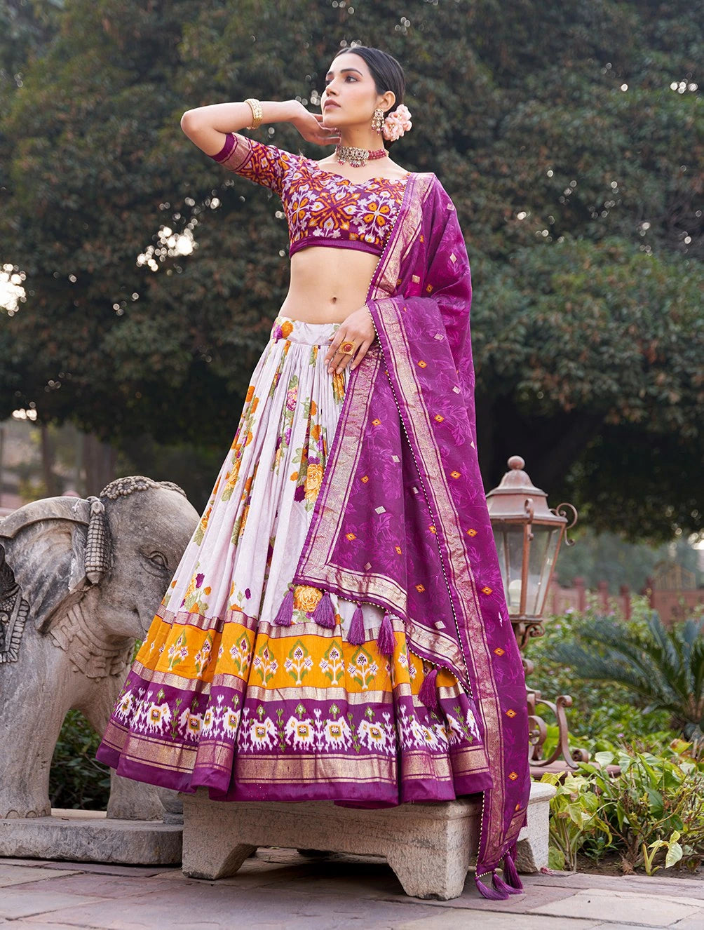 Woman in a traditional purple and white saree with floral patterns standing outdoors.
