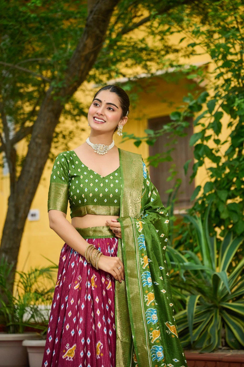 Woman in a green and pink traditional outfit standing outdoors with plants and a yellow wall in the background