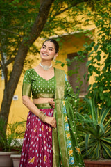 Woman in a green and pink traditional outfit standing outdoors with plants and a yellow wall in the background