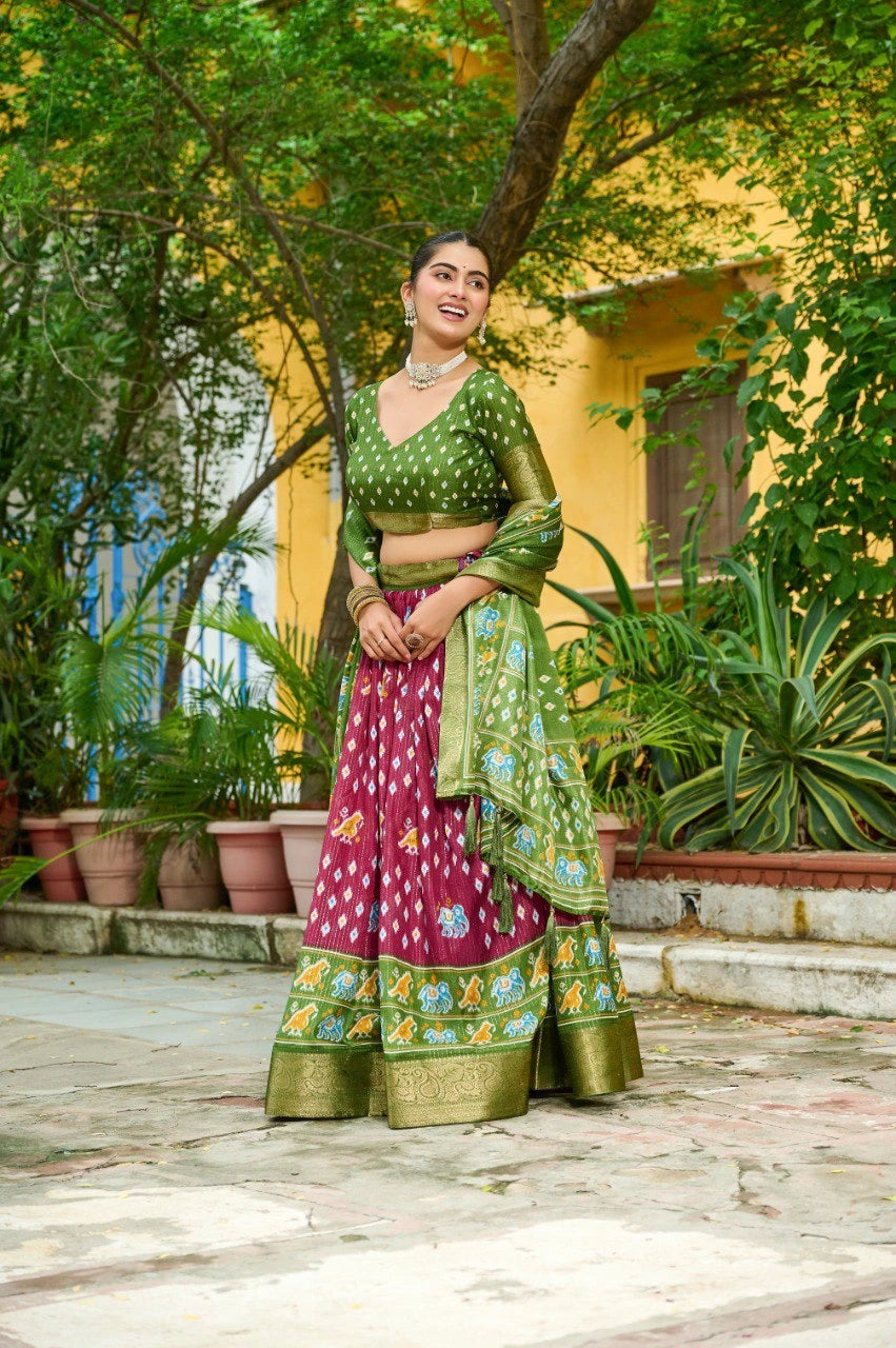 Woman in traditional green and pink saree standing outdoors with plants and a yellow building in the background
