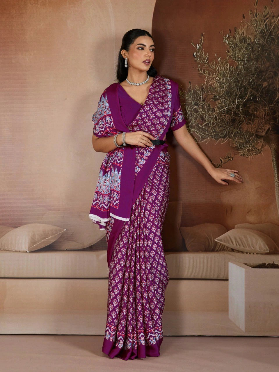 Woman in a purple saree with white patterns standing in a room with beige walls and decor.