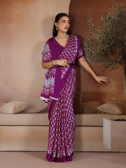 Woman in a purple saree with white patterns standing in a room with beige walls and decor.