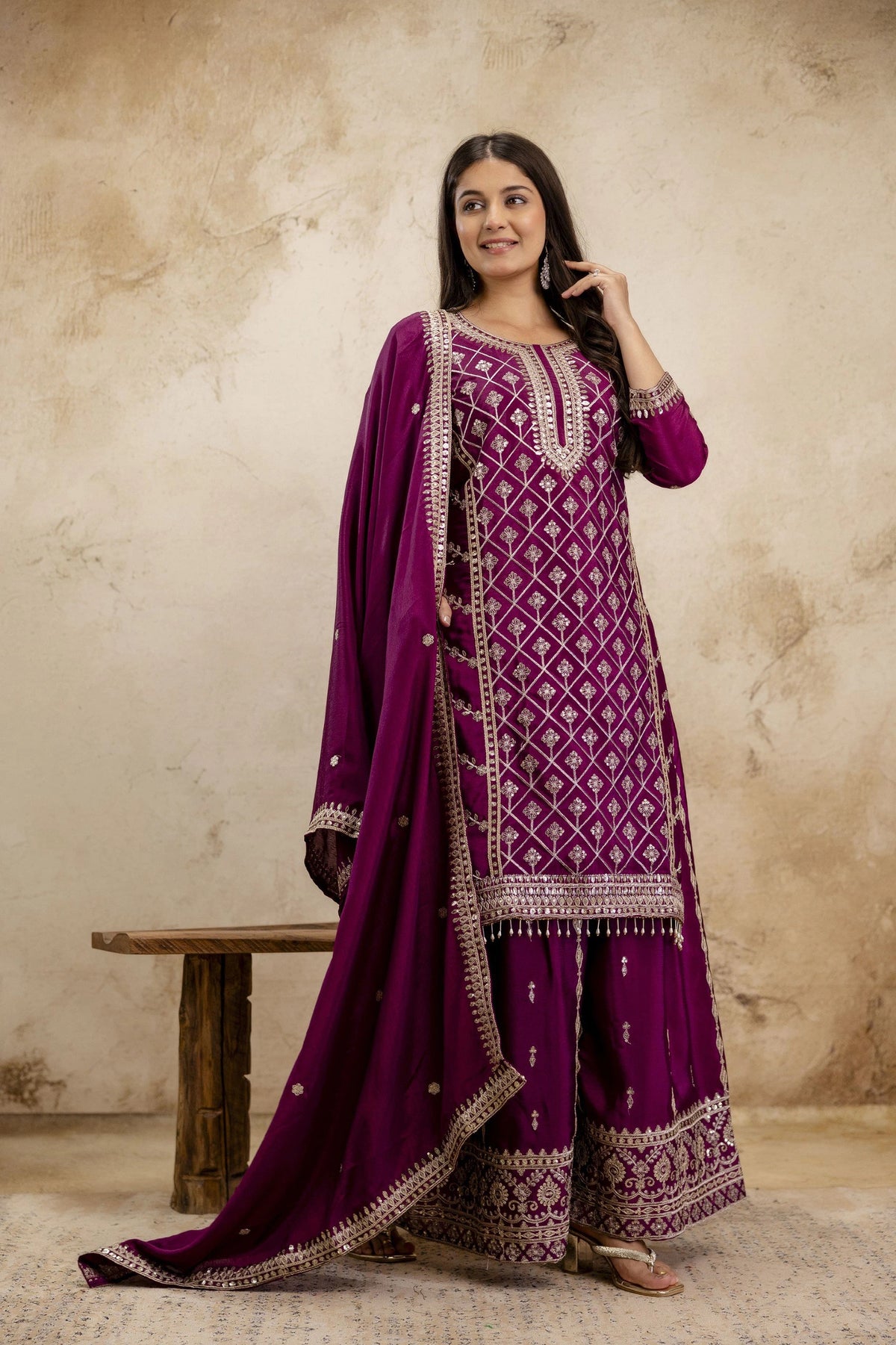 Woman wearing a purple traditional outfit with intricate patterns against a beige wall.