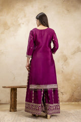Woman wearing a purple traditional outfit with intricate patterns, standing against a beige wall.