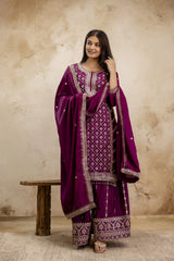 Woman wearing a traditional purple outfit with intricate patterns against a beige wall.