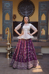 Woman in a traditional outfit with a white top and purple skirt standing in a decorated room.