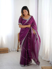 Woman wearing a purple saree with white patterns in a bright room.