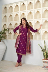 Woman in a purple traditional outfit with a patterned scarf standing against a decorative wall.
