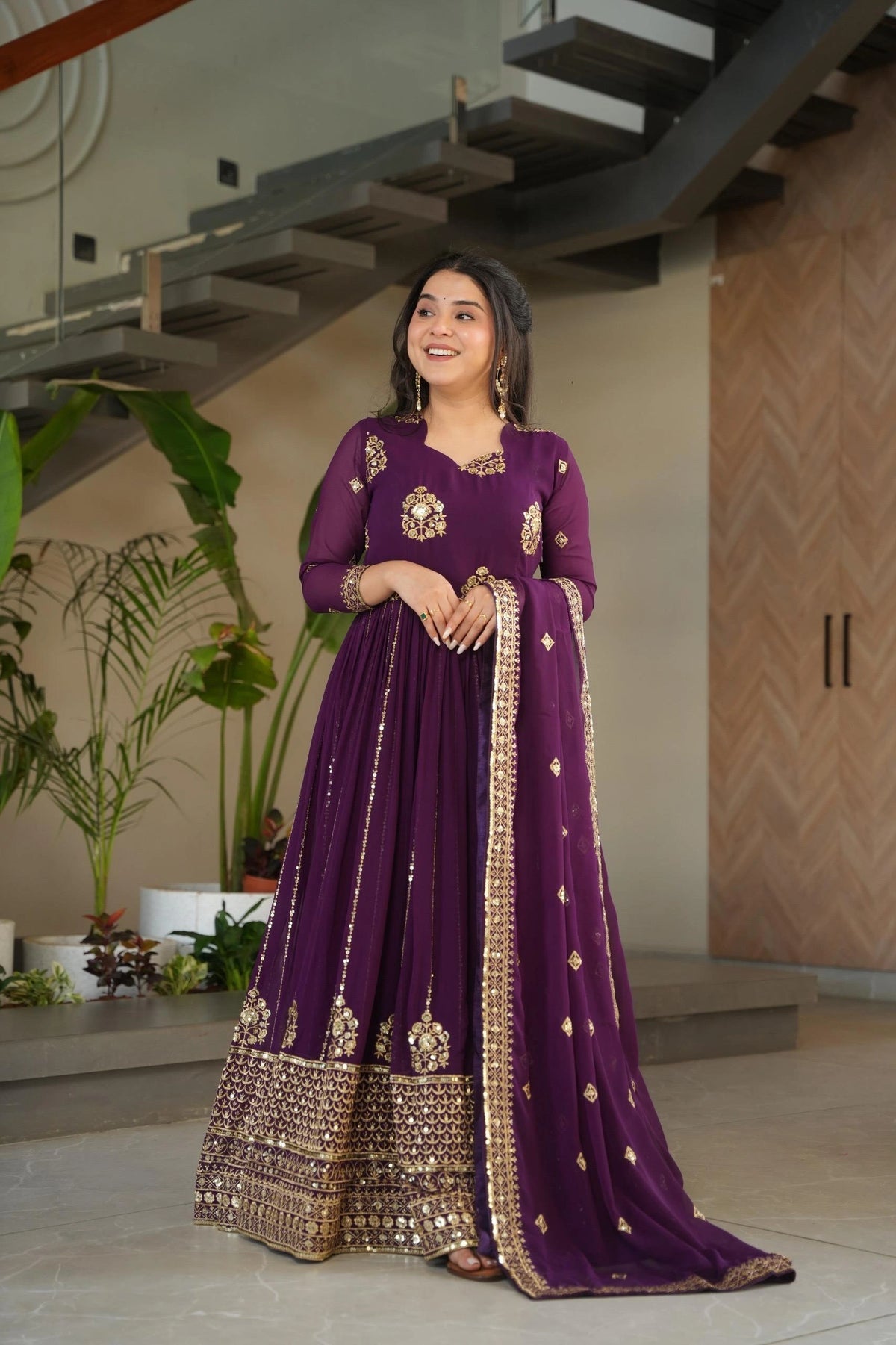 Woman in a purple traditional outfit standing indoors with plants and architectural elements in the background
