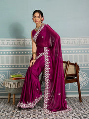 Woman in a purple saree with white patterns standing in a room with decorative walls and furniture.