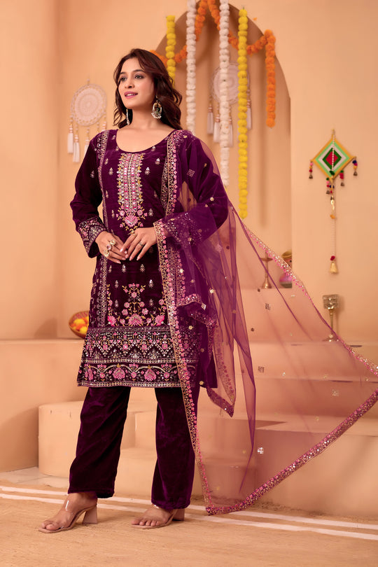 Woman wearing a purple embroidered traditional outfit with a sheer dupatta in a decorated room.