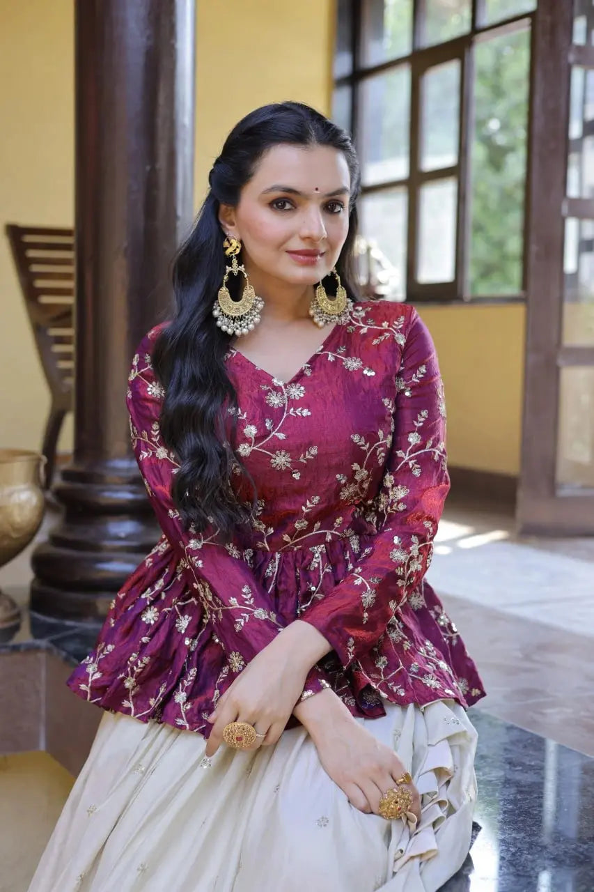 Woman in a traditional outfit with a floral top and white skirt, sitting indoors.