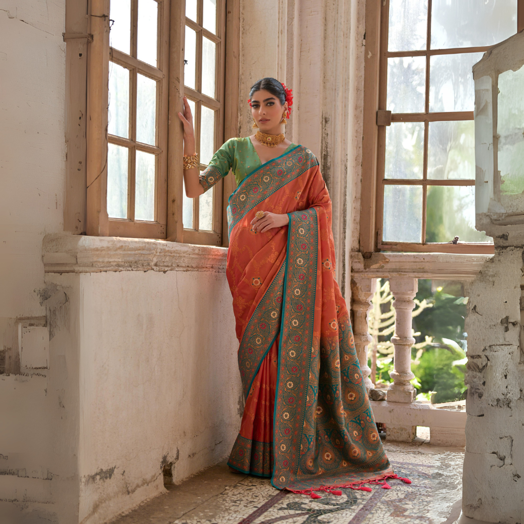 A woman models a vibrant Coral Orange Banarasi Silk Saree with all-over gold Zari weaving. The saree has a wide, ornate Rama Blue/Teal border featuring traditional Zari weaving. She wears a light green blouse and heavy gold Kundan jewelry, posing by a large antique window.