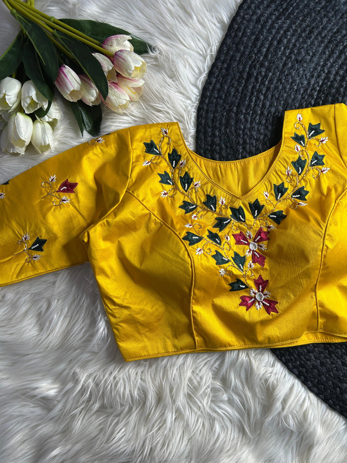 A vibrant yellow silk blouse with a deep round neckline, featuring intricate hand-embroidery along the neckline and on the sleeves. The design showcases gold-toned beaded vines, dark green leaves, and maroon floral motifs with white beaded centers. The blouse is laid flat on a white faux-fur surface next to a gray woven mat and a bunch of white and pink tulips.