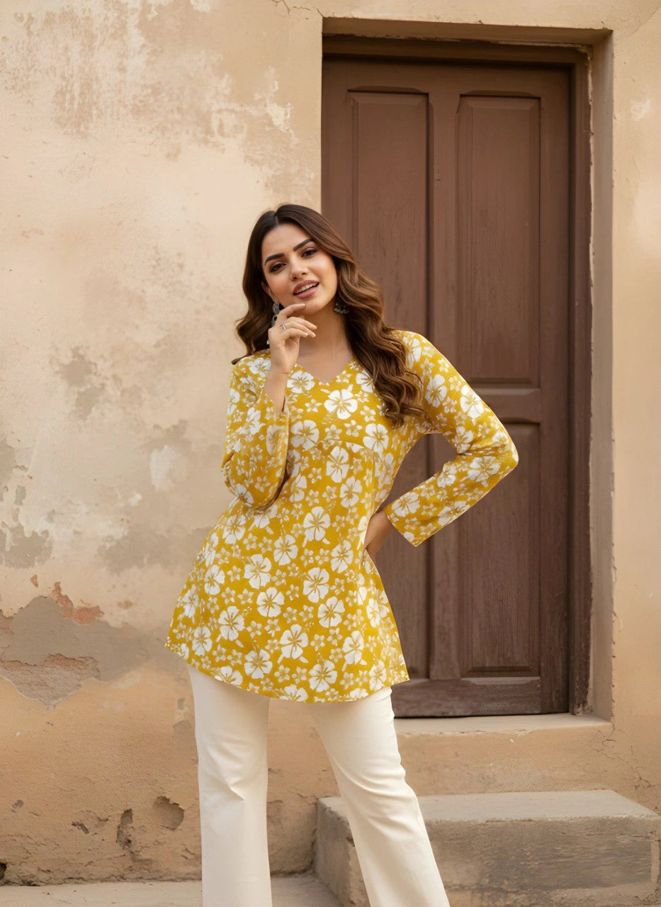 The woman poses with her hand on her hip, highlighting the peplum-style silhouette of the short kurti. The vibrant white floral pattern is clearly visible against the mustard fabric.