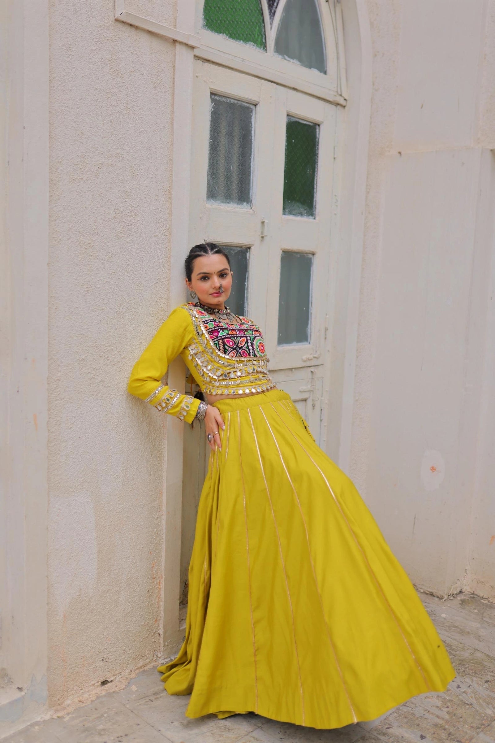 Woman in a yellow traditional outfit standing against a white wall with a window.