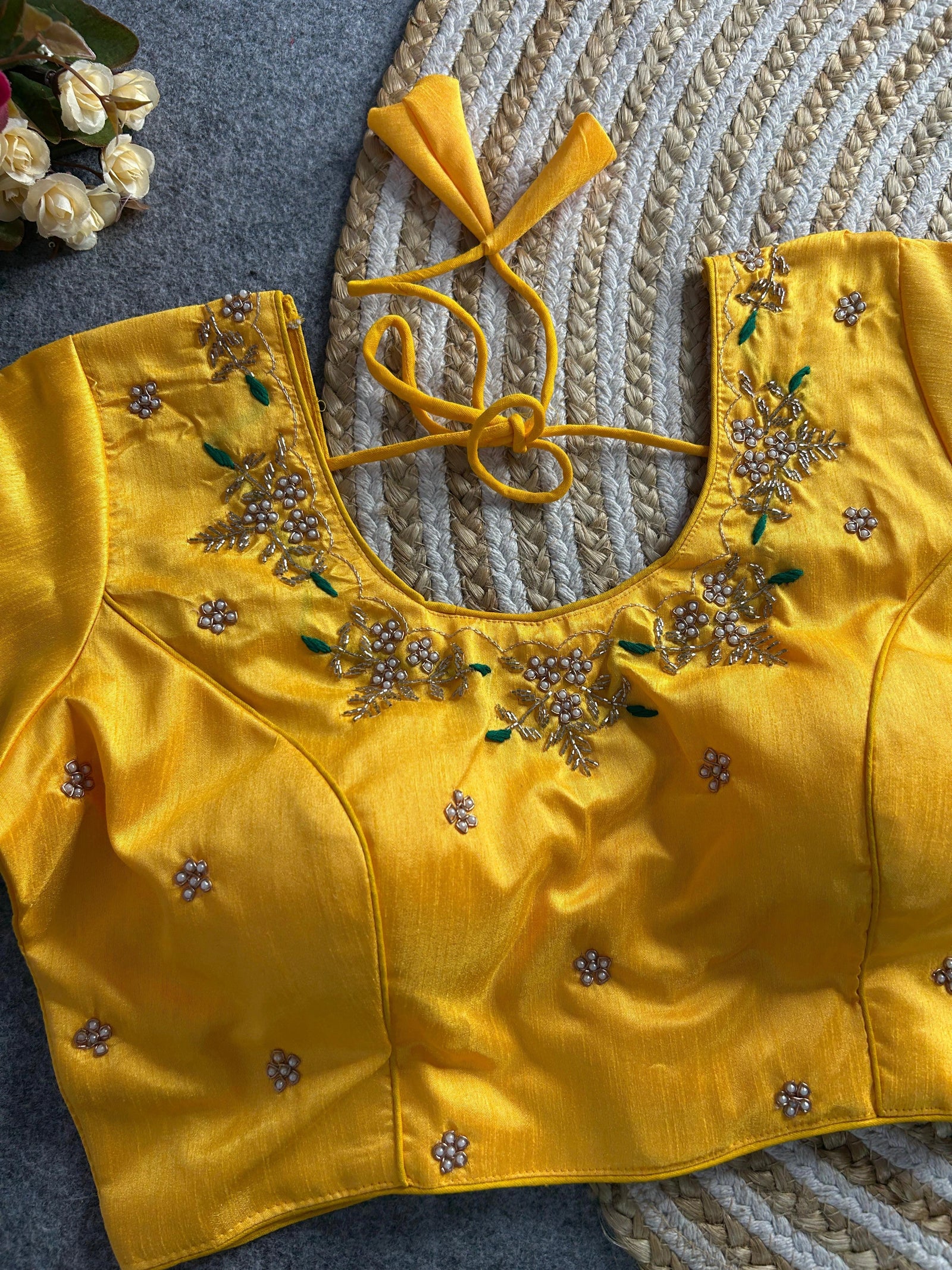 The front of the yellow silk blouse features a curved V-neckline with matching pearl and gold floral handwork. The bodice is tailored with princess-line seams and is adorned with scattered embroidered pearl motifs. The blouse is displayed on a grey textured surface next to a bouquet of maroon and cream flowers.