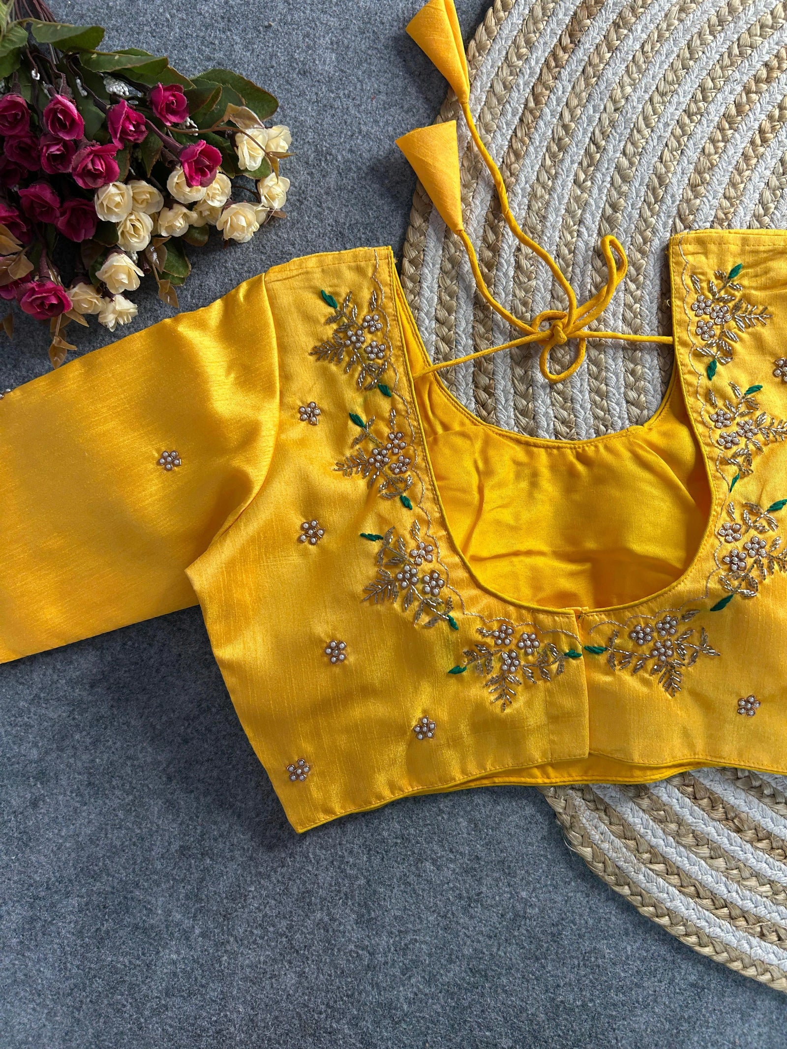 A bright yellow raw silk blouse with a deep U-neckline at the back. The edges are decorated with intricate gold and pearl floral embroidery featuring small green leaf accents. It includes matching yellow tie-back cords with double-layered triangular tassels, and small pearl buttis are scattered across the fabric.