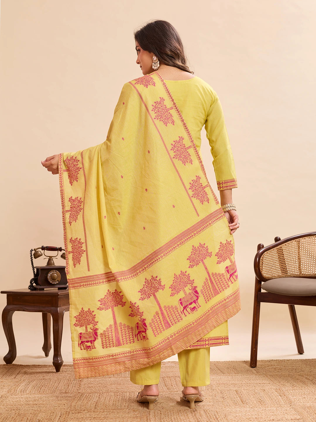 Woman wearing a yellow ethnic suit with pink traditional prints, shown from the back, holding a matching dupatta with detailed border patterns and motifs, styled with heels in a minimal indoor setting.