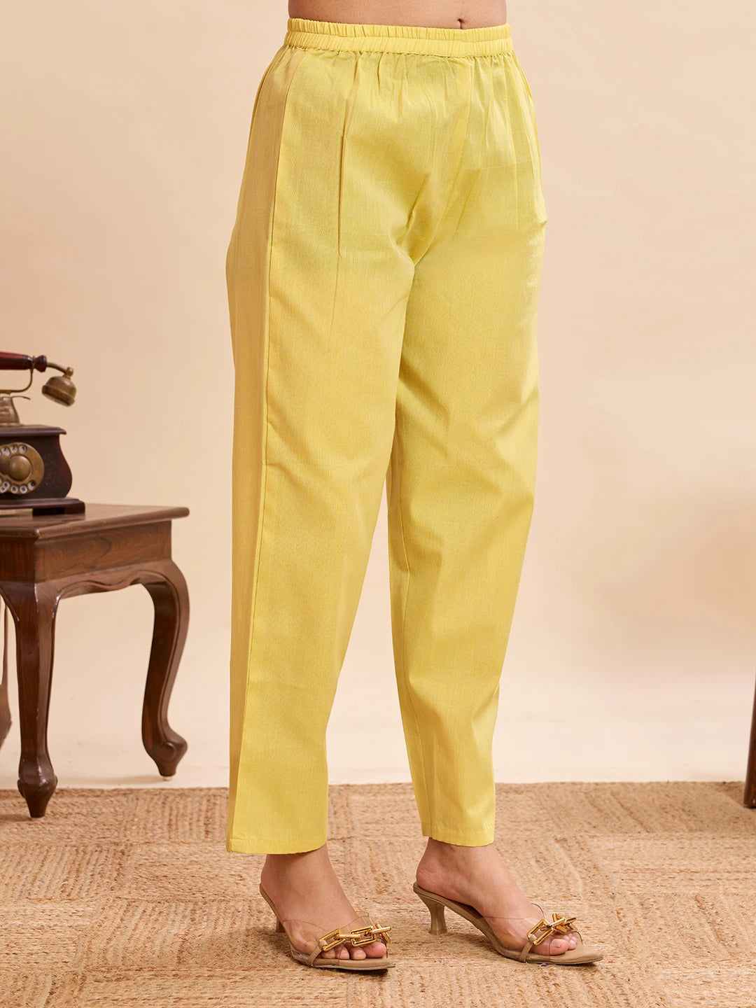 Yellow straight-fit pants with elastic waistband, paired with transparent heeled sandals, styled against a neutral background with a vintage side table.