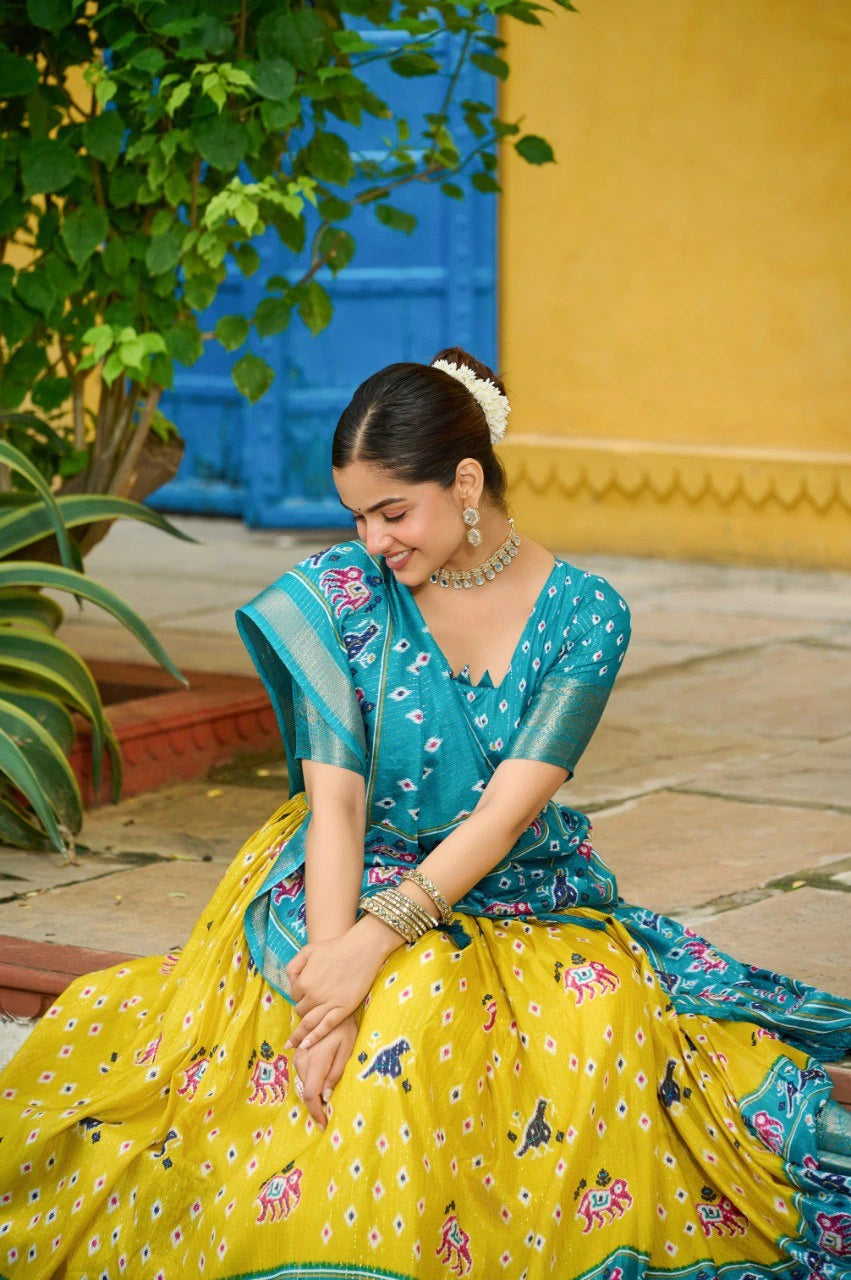 Woman in a traditional yellow and blue saree sitting outdoors with greenery and a yellow wall in the background.