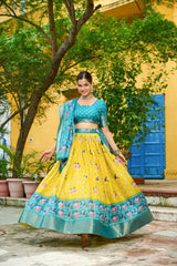 Woman in a yellow and green traditional outfit standing outdoors with greenery and a yellow wall in the background.