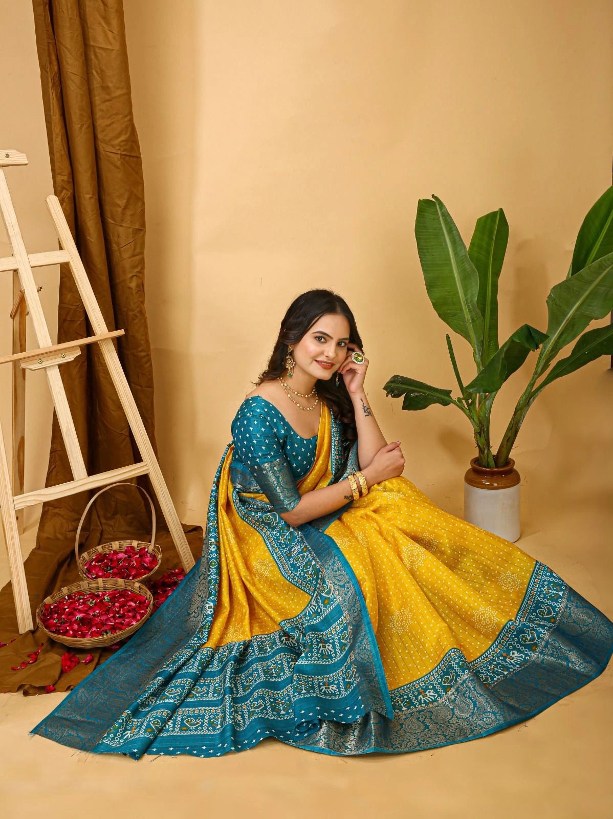 Woman in a yellow and blue saree sitting on the floor with a plant and basket in the background.