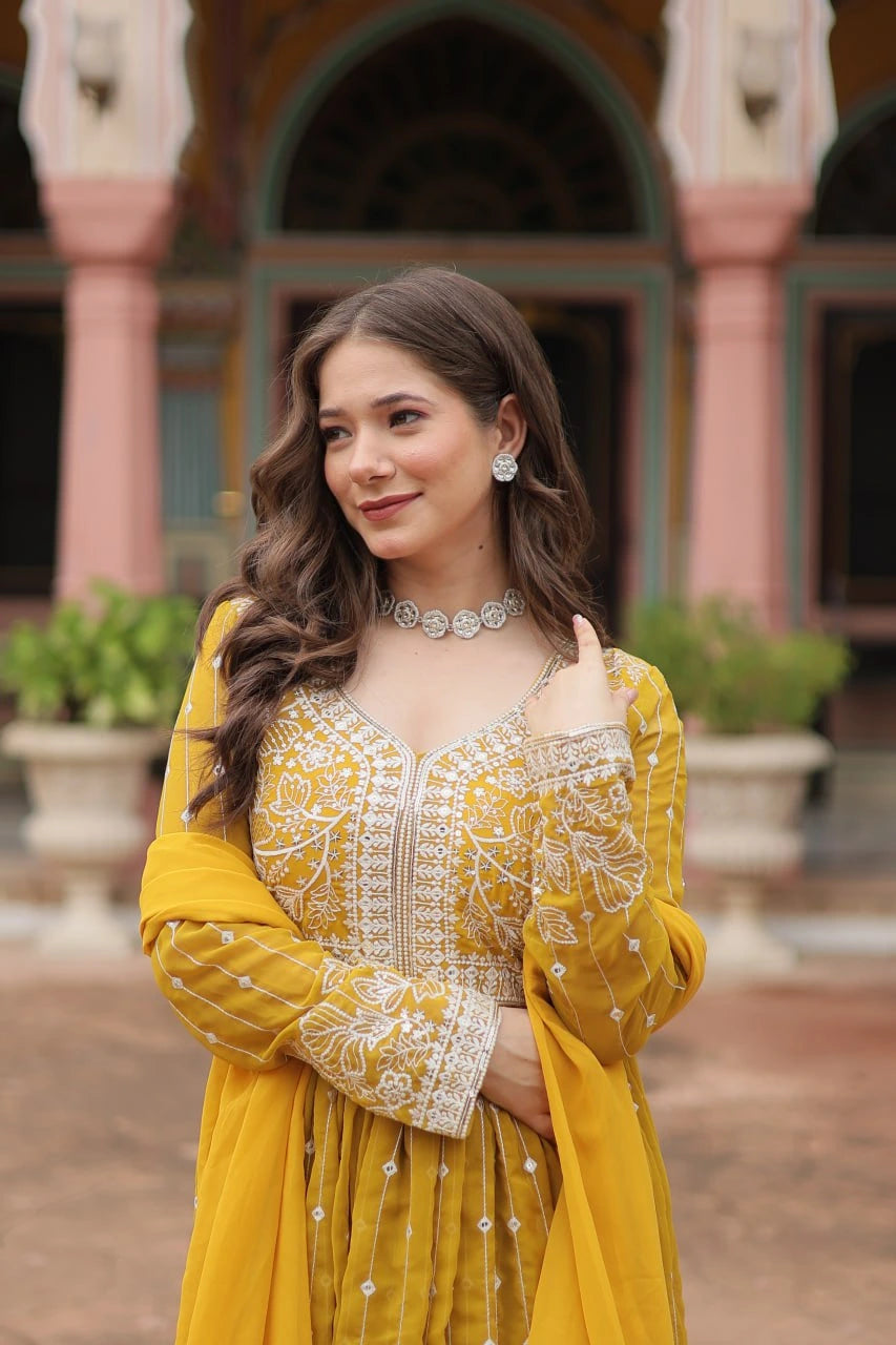Woman in a yellow traditional outfit with intricate patterns, standing in front of an architectural background.