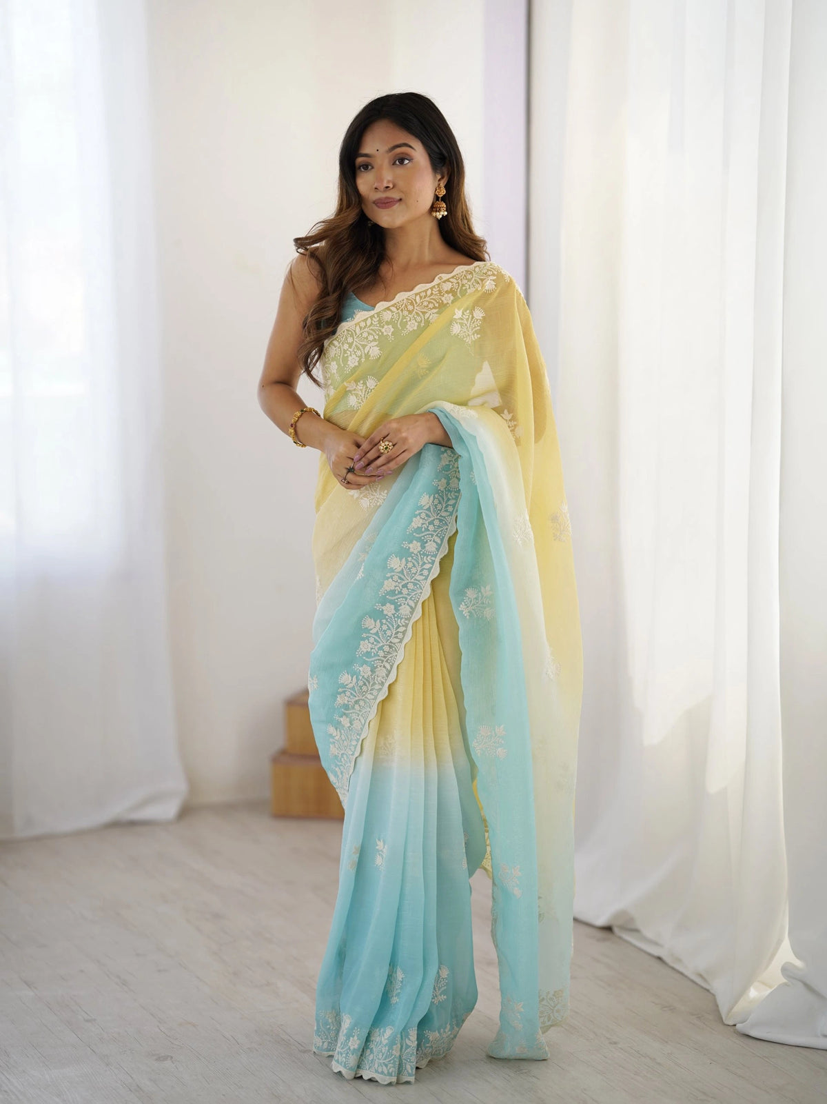 Woman wearing a yellow and blue saree with white lace details against a white curtain background
