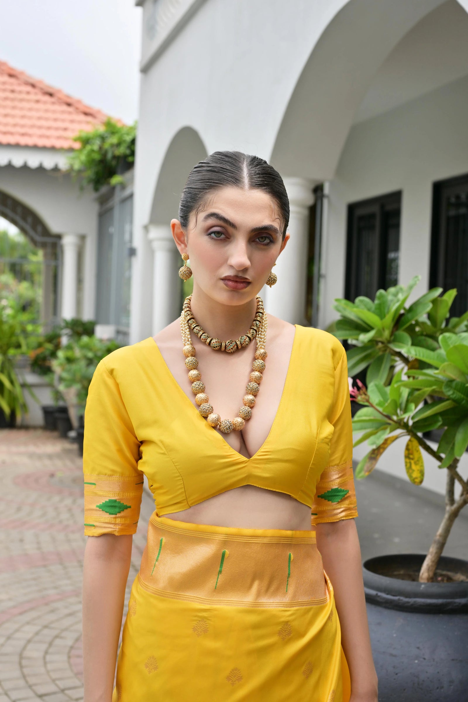 Woman in a yellow traditional outfit standing outdoors with greenery and a building in the background