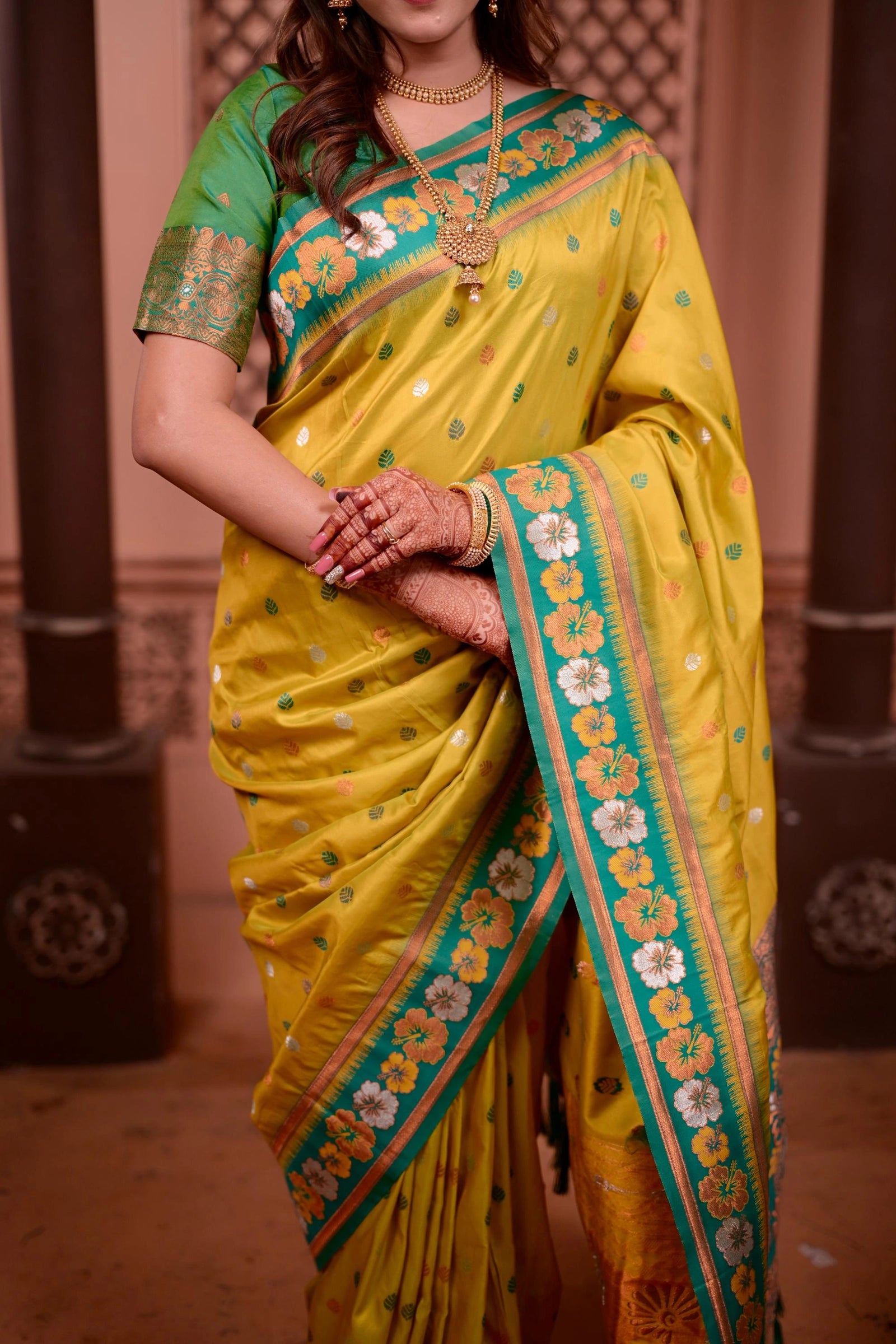 Woman wearing a yellow saree with green border and floral patterns indoors.