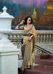 Woman in a traditional beige saree with intricate patterns standing on steps with a decorative wall in the background.