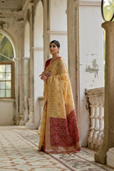 Woman in a yellow saree with a red border standing in an ornate room.