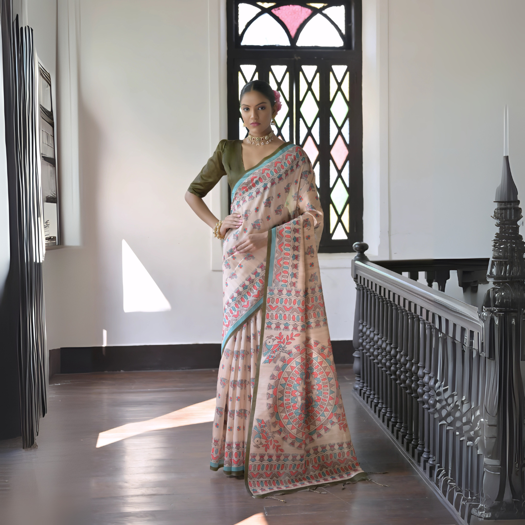 A woman models an elegant Beige Soft Tussar Silk Saree featuring an all-over Madhubani-style print in teal, coral, and green. The saree has a dark green and aqua border. She wears a contrasting olive green short-sleeve blouse and a gold Kundan choker necklace, posing indoors next to a dark wooden banister.