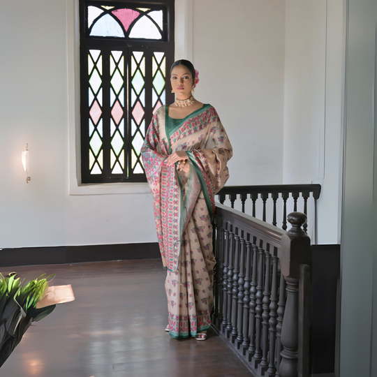 A woman models an elegant Off-White Soft Banarasi Silk Saree. The saree features a wide, prominent border and pallu with rich gold Zari weaving/print in shades of orange, peach, and green. She wears a coral sleeveless blouse and accessorizes with a gold Kundan choker necklace, large earrings, and a distinct dome-shaped handbag, posing indoors against a deep red backdrop.