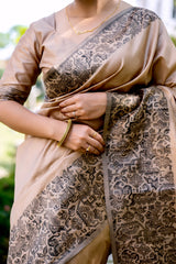 A woman models a luxurious Beige/Taupe Raw Silk Saree. The saree features a wide border and pallu with dense, intricate dark brown/black Zari weaving in a Kalamkari-style floral and paisley pattern. She wears a matching short-sleeve beige blouse and traditional gold jewelry, posing outdoors on a stone pathway.
