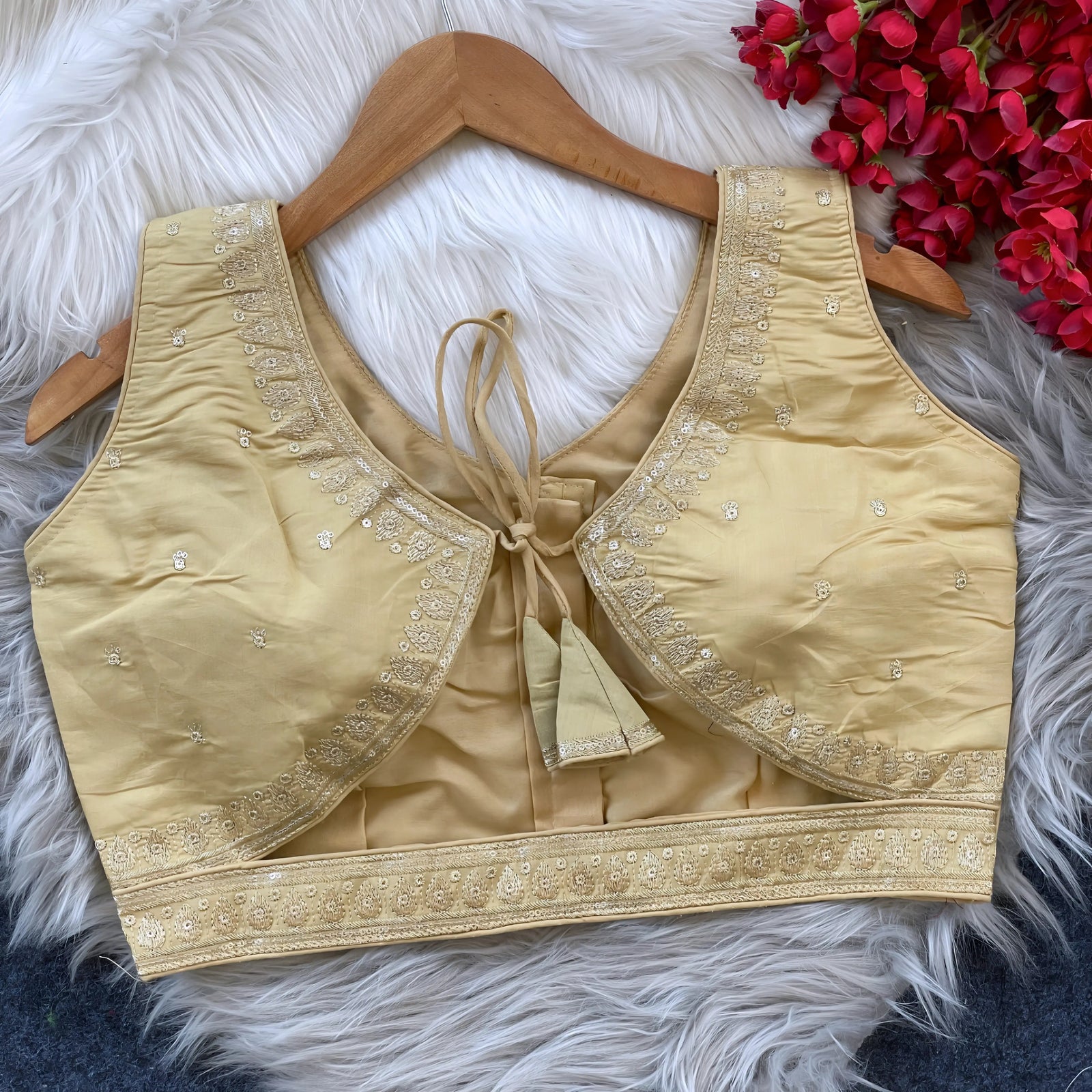 Beige padded blouse with delicate gold embroidery along the neckline and hem, featuring small sequin floral motifs, displayed on a wooden hanger over a white fur surface with red artificial flowers in the background.