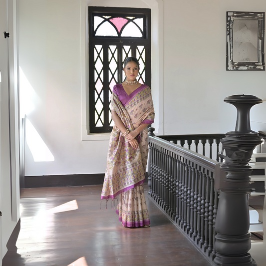 Woman wearing a beige silk saree with intricate floral motifs and a purple border, accessorized with a choker necklace and bangles, standing in a sunlit vintage interior with woA woman models an elegant Beige Soft Tussar Silk Saree. The saree features an all-over Madhubani-style print of traditional motifs primarily in soft purple/lilac and yellow/green. It is framed by a vibrant deep purple/magenta border. She wears a matching purple sleeoden flooring, black railing, and a window with stained glass panels.
