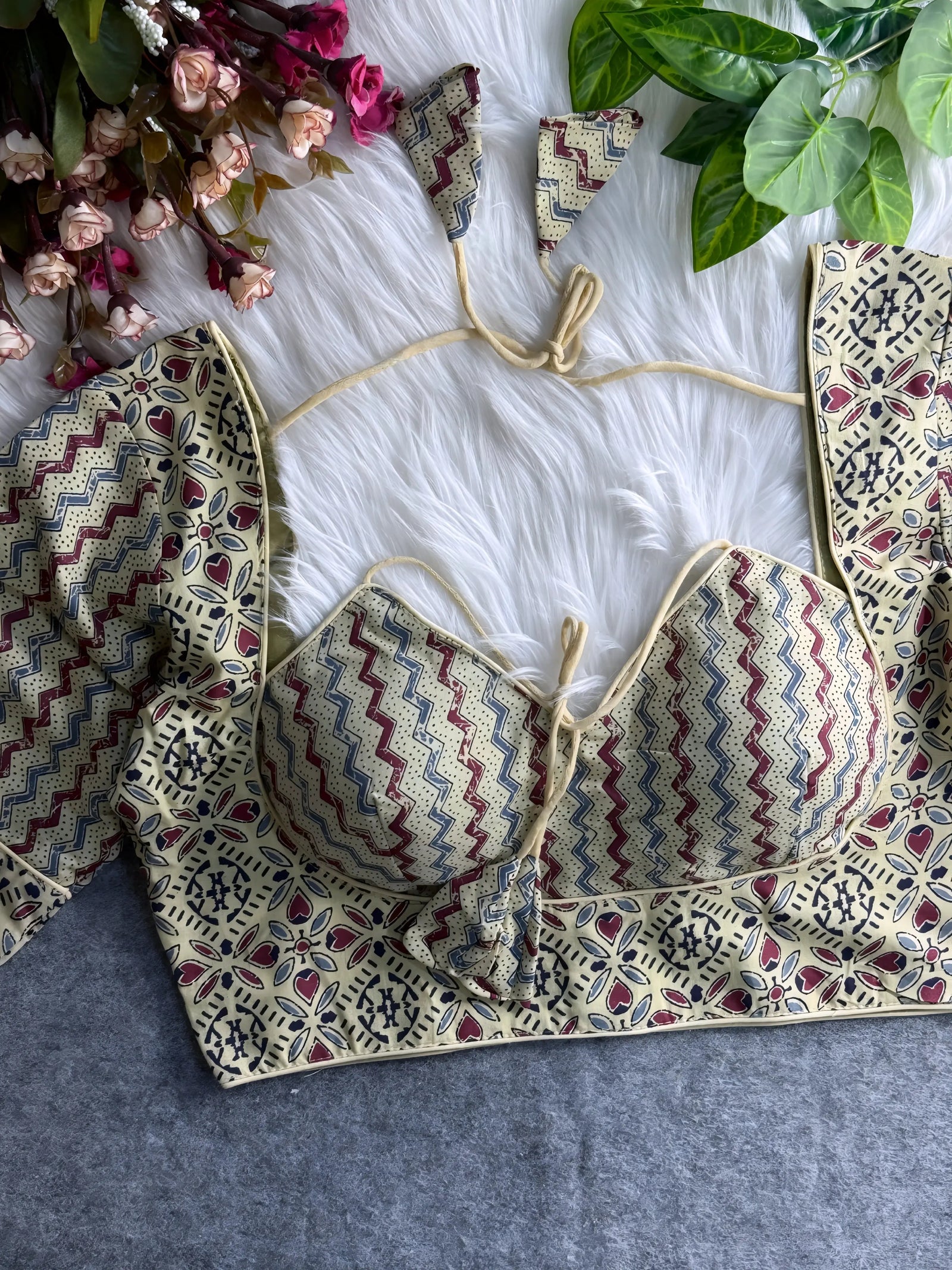 Beige padded blouse with maroon and navy geometric and floral patterns, displayed on a white fur surface with artificial flowers and green leaves in the background.