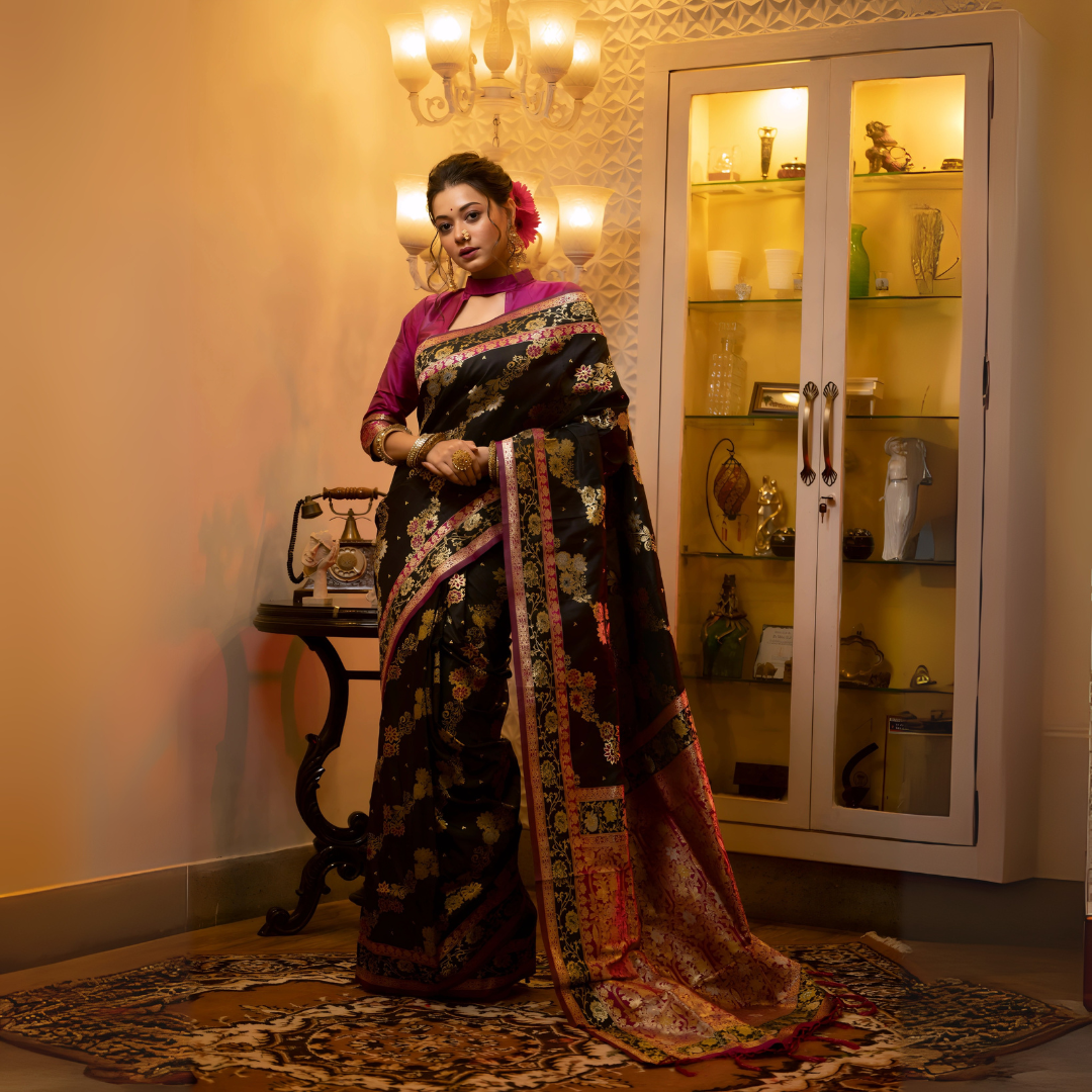 A woman models a luxurious Black Soft Banarasi Silk Saree. The saree features dense, multi-colored Zari weaving of gold and pink florals, and a wide pallu with heavy gold and Fuchsia Pink accents. She wears a high-neck magenta blouse and gold jewelry, posing in a dramatically lit antique setting.