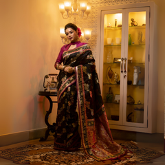 A woman models a luxurious Black Soft Banarasi Silk Saree. The saree features dense, multi-colored Zari weaving of gold and pink florals, and a wide pallu with heavy gold and Fuchsia Pink accents. She wears a high-neck magenta blouse and gold jewelry, posing in a dramatically lit antique setting.
