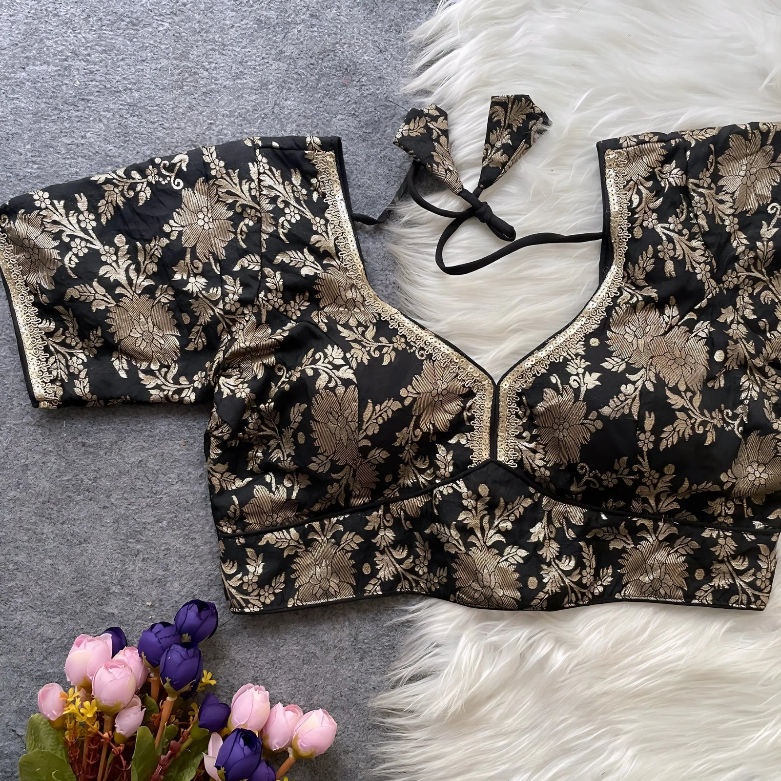 Black brocade blouse featuring intricate gold floral zari embroidery and decorative trim along the neckline and sleeves, displayed flat on a grey surface with a white faux fur accent and a bunch of artificial pink and purple flowers in the corner, showcasing its festive ethnic design and elegant craftsmanship.