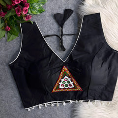 Black padded blouse with white piping, featuring a central embroidered triangle motif with mirror work and shell detailing along the hem, displayed on a grey surface with artificial pink flowers and white fur in the background.