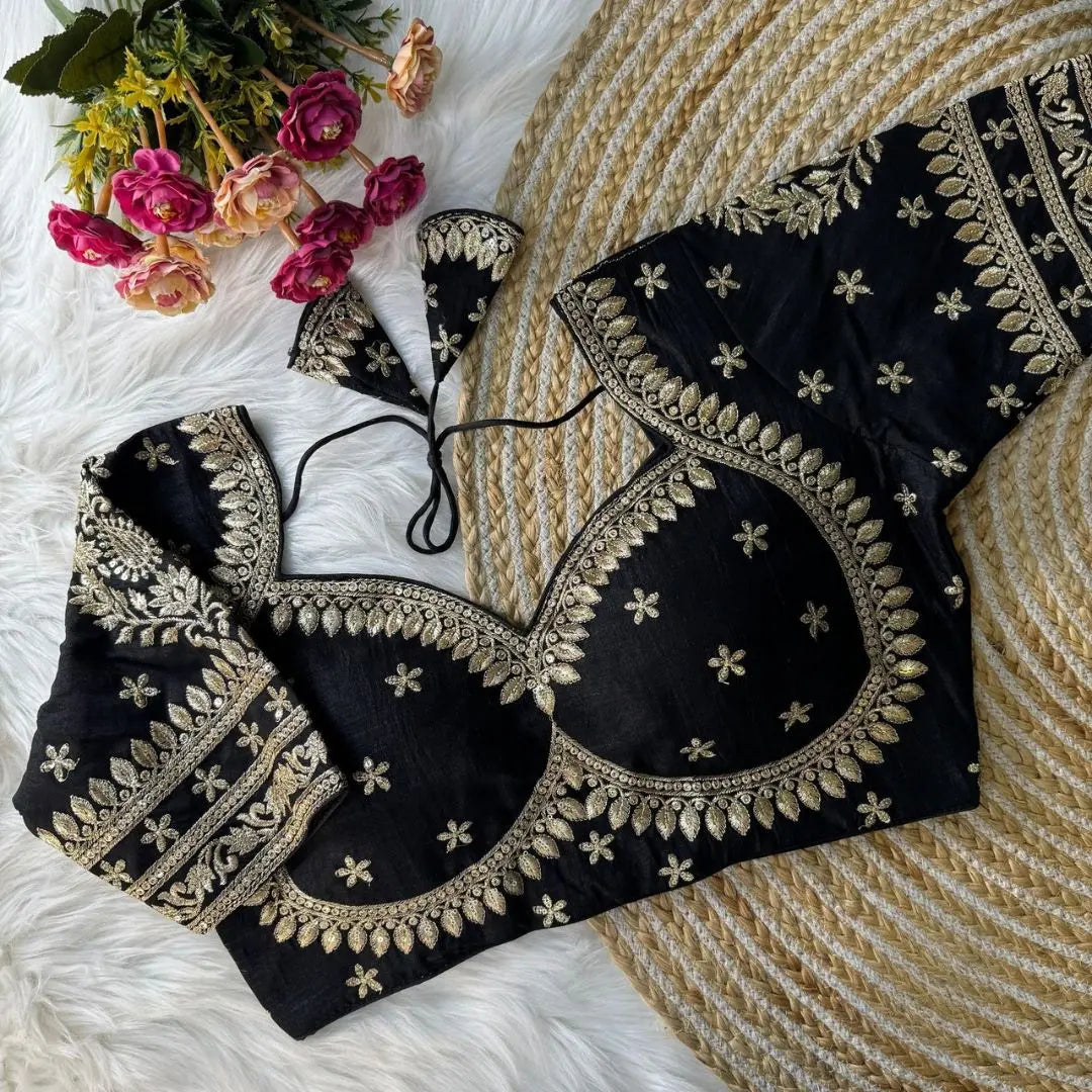 Black embroidered silk blouse with intricate gold zari floral motifs and a tie-back design, displayed on a white fur background with a bunch of pink and peach flowers in the corner