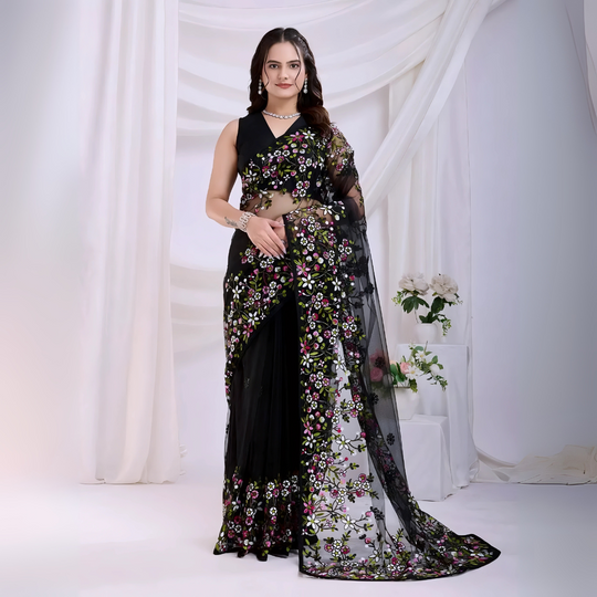 A woman models a dramatic Black Soft Net Saree. The sheer fabric is densely covered in vibrant all-over floral embroidery featuring rich pink, white, and green threadwork. She wears a matching black sleeveless blouse and accessorizes with sophisticated silver/diamond jewelry, posing indoors against sheer white curtains.