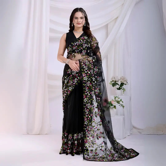 A woman models a dramatic Black Soft Net Saree. The sheer fabric is densely covered in vibrant all-over floral embroidery featuring rich pink, white, and green threadwork. She wears a matching black sleeveless blouse and accessorizes with sophisticated silver/diamond jewelry, posing indoors against sheer white curtains.
