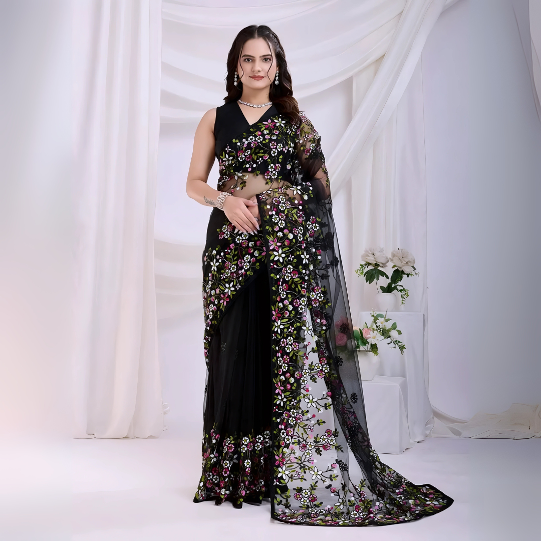 A woman models a dramatic Black Soft Net Saree. The sheer fabric is densely covered in vibrant all-over floral embroidery featuring rich pink, white, and green threadwork. She wears a matching black sleeveless blouse and accessorizes with sophisticated silver/diamond jewelry, posing indoors against sheer white curtains.