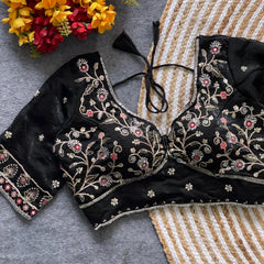 Black padded blouse with intricate floral embroidery in gold and red threads, featuring a tie-back detail, displayed on a grey surface with artificial yellow and red flowers and a woven mat in the background.
