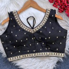 Black padded blouse with intricate gold embroidery along the neckline and hem, featuring small gold embroidered motifs, displayed on a wooden hanger over a white fur surface with red artificial flowers in the background.
