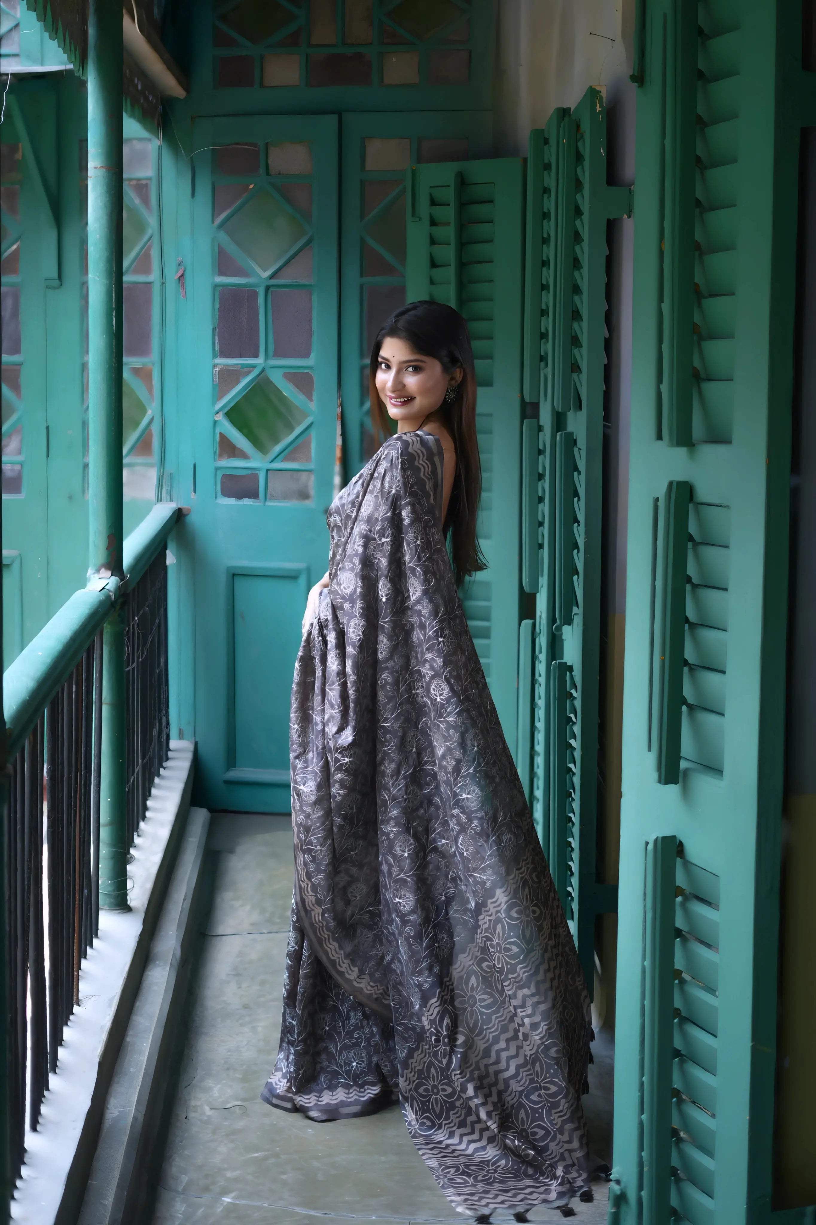A woman models an elegant Black Tussar Silk Saree. The saree features dense all-over floral embroidery/weaving in a shimmering metallic silver thread. She wears a black sheer-sleeve blouse and heavy oxidized silver jewelry, including a large ring and stacked bangles, posing in a doorway framed by vibrant teal shutters.